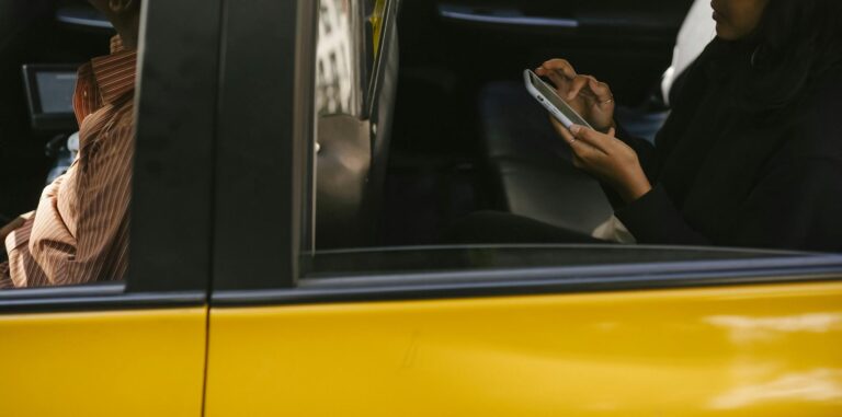 Side view of unrecognizable passenger browsing smartphone while sitting on backseat of yellow cab with anonymous driver during ride